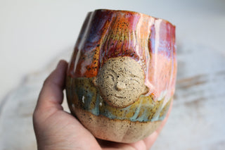 a hand holding a ceramic cup with a face on it